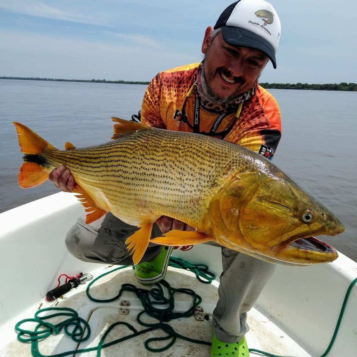 Un pescador sostiene con las dos manos un dorado grande sobre la lancha, con el río Uruguay de fondo