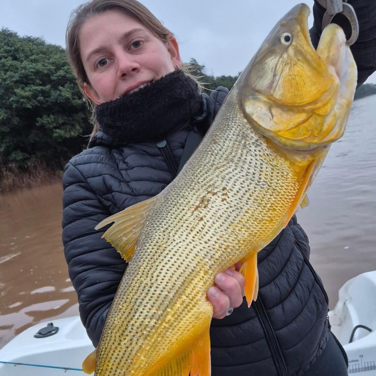 Una pescadora sostiene un dorado de gran porte en una mañana nublada sobre el río