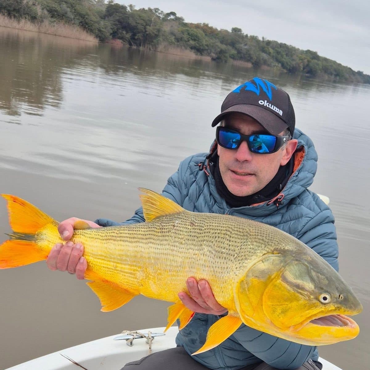 Un pescador con el dorado en las manos, con la costa arbolada del río Uruguay detrás