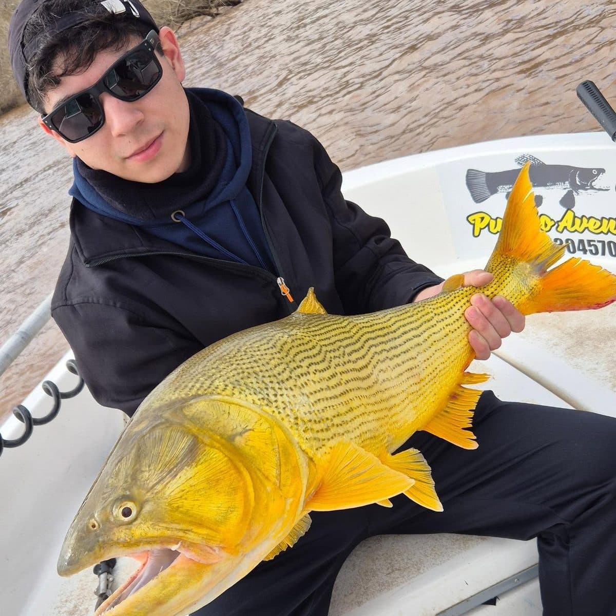Un pescador joven sostiene un dorado grande sobre la lancha, con el río de fondo