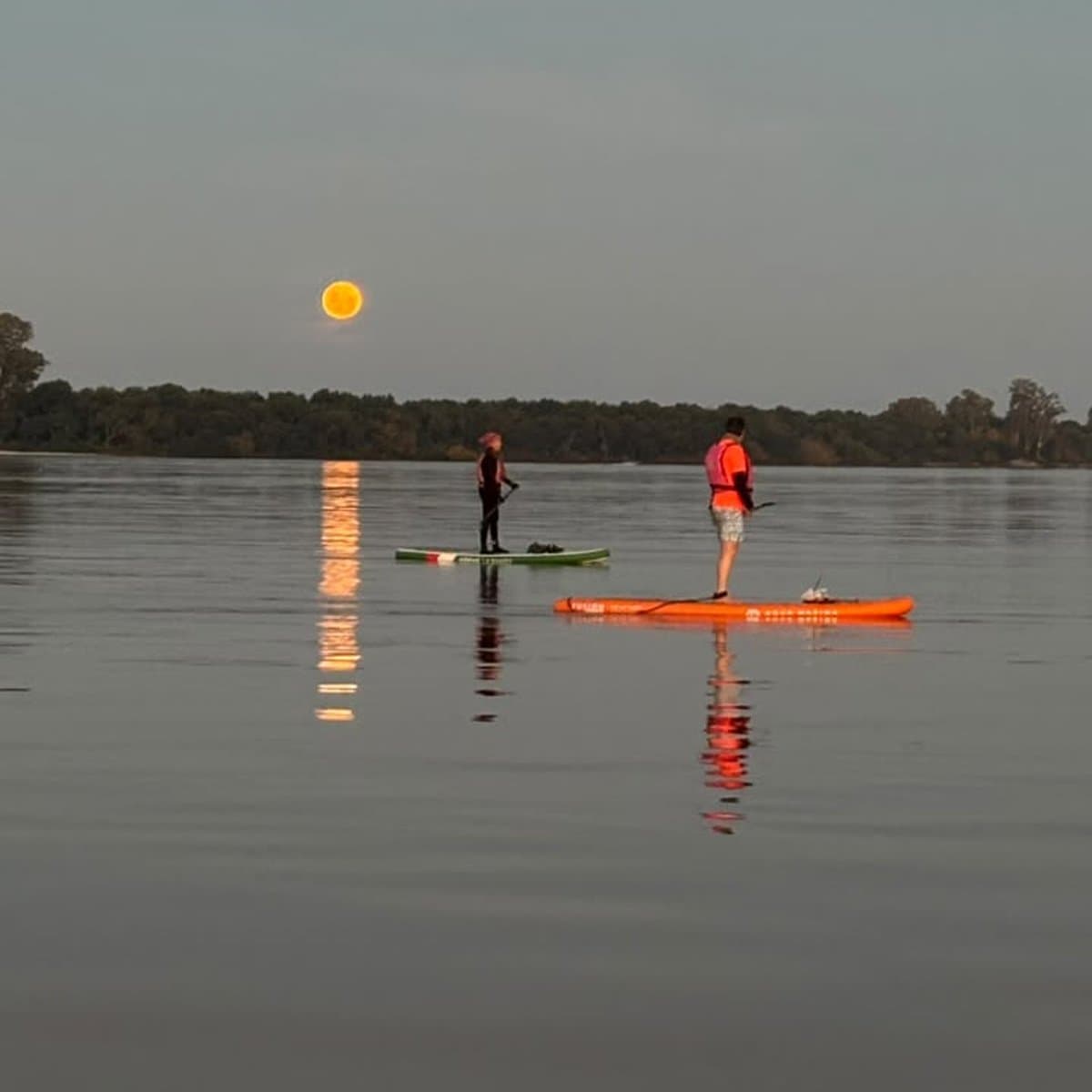 Dos remeros de pie en sus tablas al anochecer, con la luna naranja reflejándose en una franja larga sobre el agua