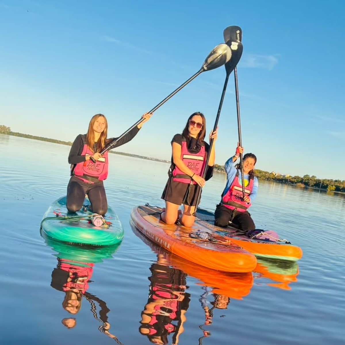 Tres mujeres arrodilladas en tablas de colores levantan los remos y los cruzan, con el río liso y el cielo despejado detrás