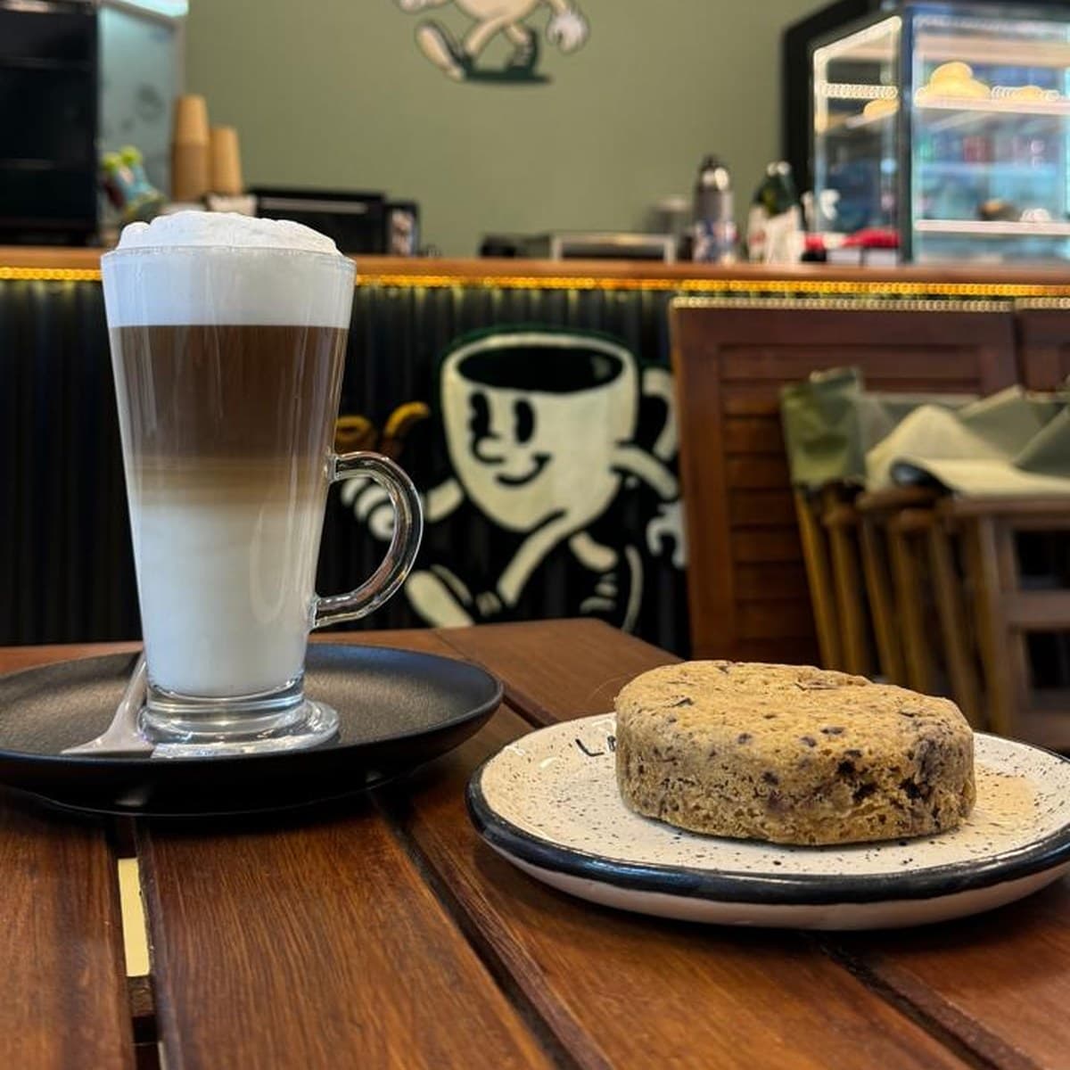 Un latte macchiato en vaso alto junto a una cookie, sobre una mesa de madera, con el mostrador y el logo del pocillo pintado al fondo