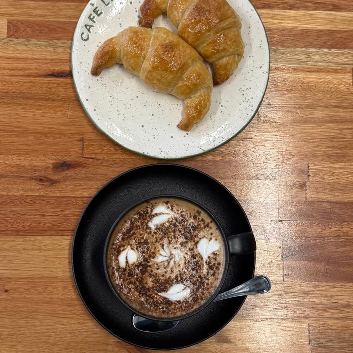 Dos medialunas en un plato con el nombre Café La Taba y un capuchino con cacao espolvoreado, vistos desde arriba sobre una mesa de madera