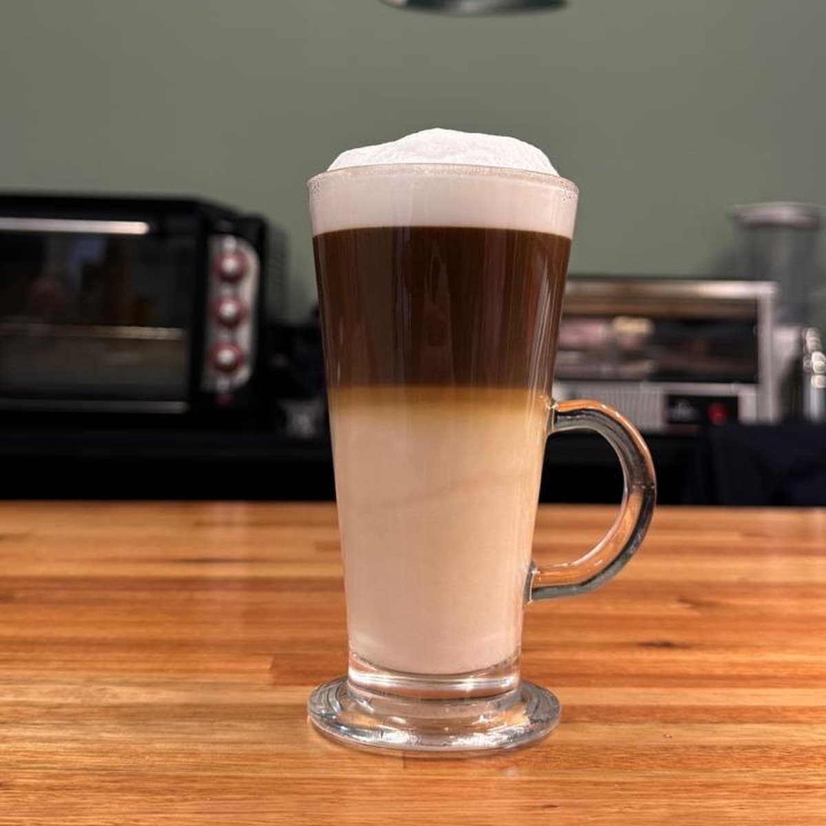 Un latte macchiato en vaso alto sobre la barra de madera, con la pared verde del salón detrás