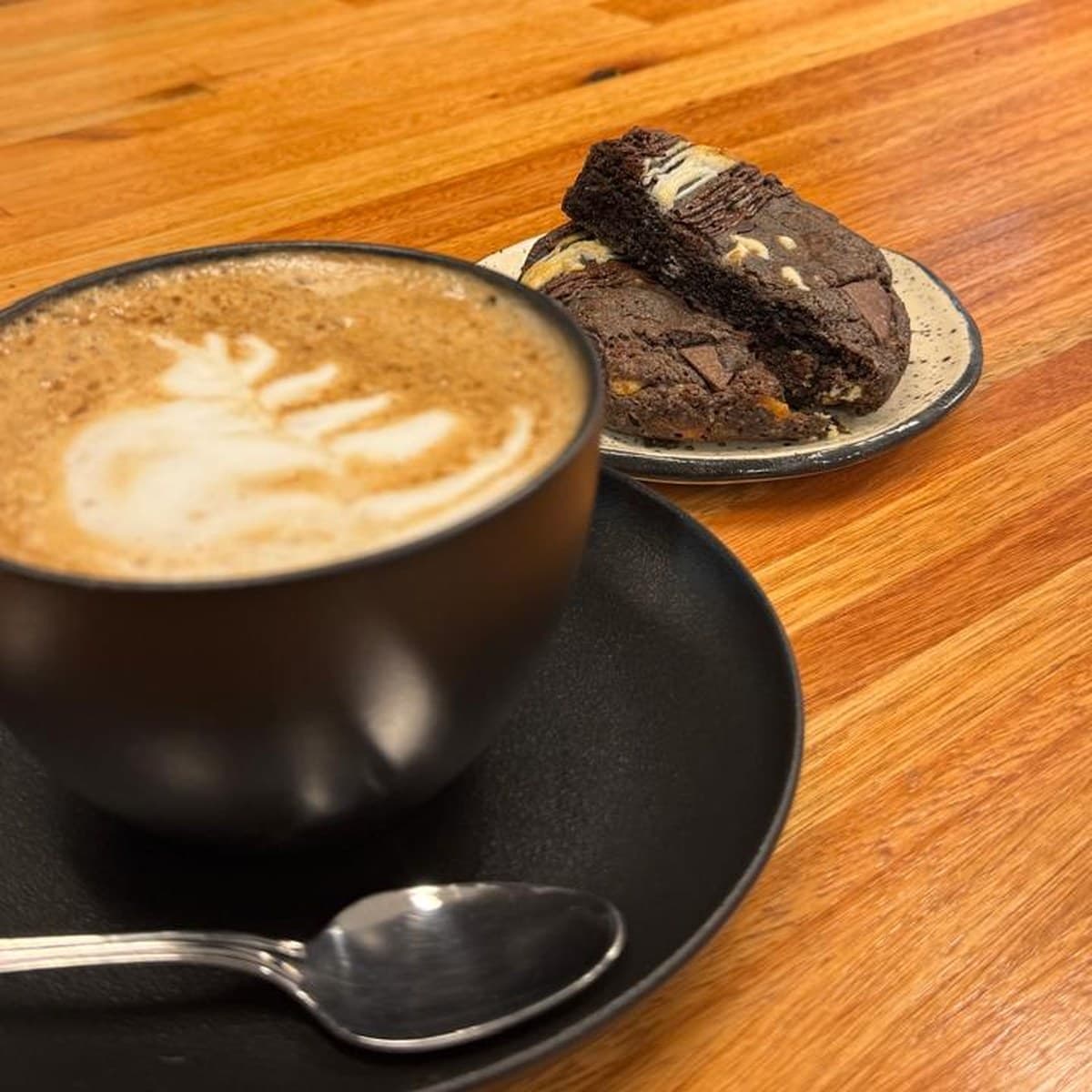 Un capuchino con arte en la leche en taza negra y un brownie con chocolate en un plato, sobre la barra de madera
