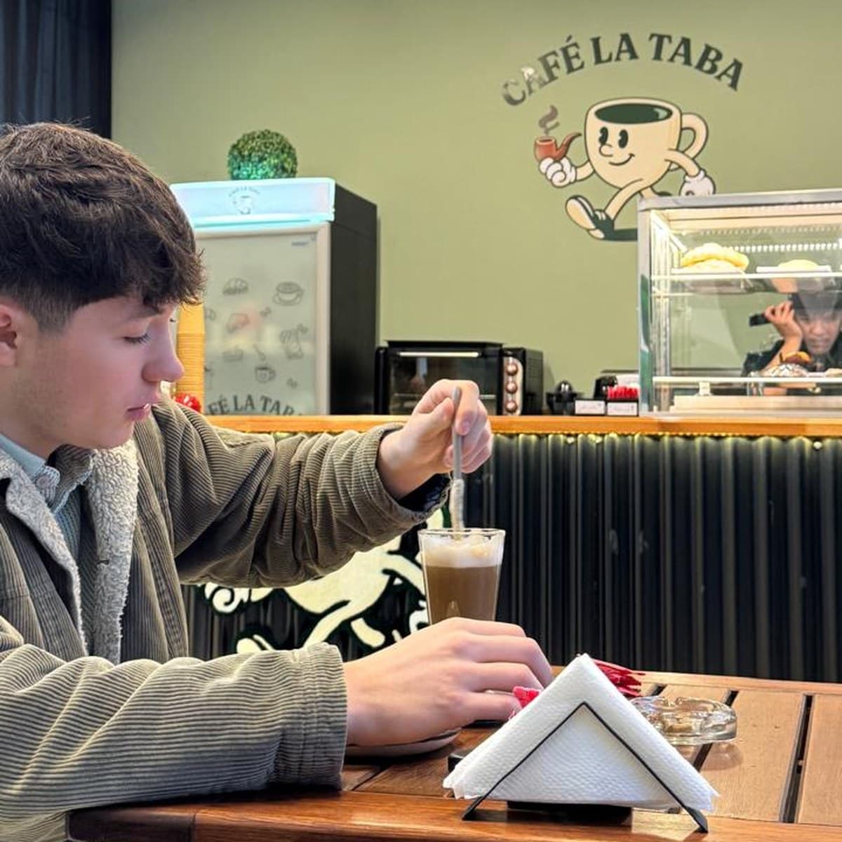 Un cliente revolviendo un café con leche en vaso alto en una mesa del salón, con el mostrador, la vitrina de facturas y el logo del pocillo pintado en la pared verde al fondo