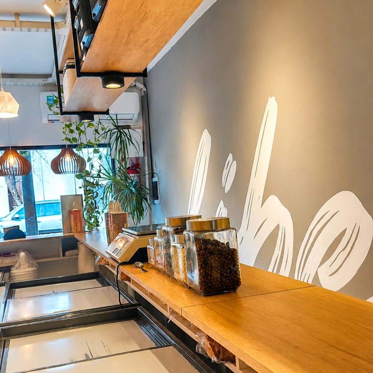 La barra de madera junto a las cubas de helado, con frascos de toppings y el lettering blanco del local pintado en la pared gris