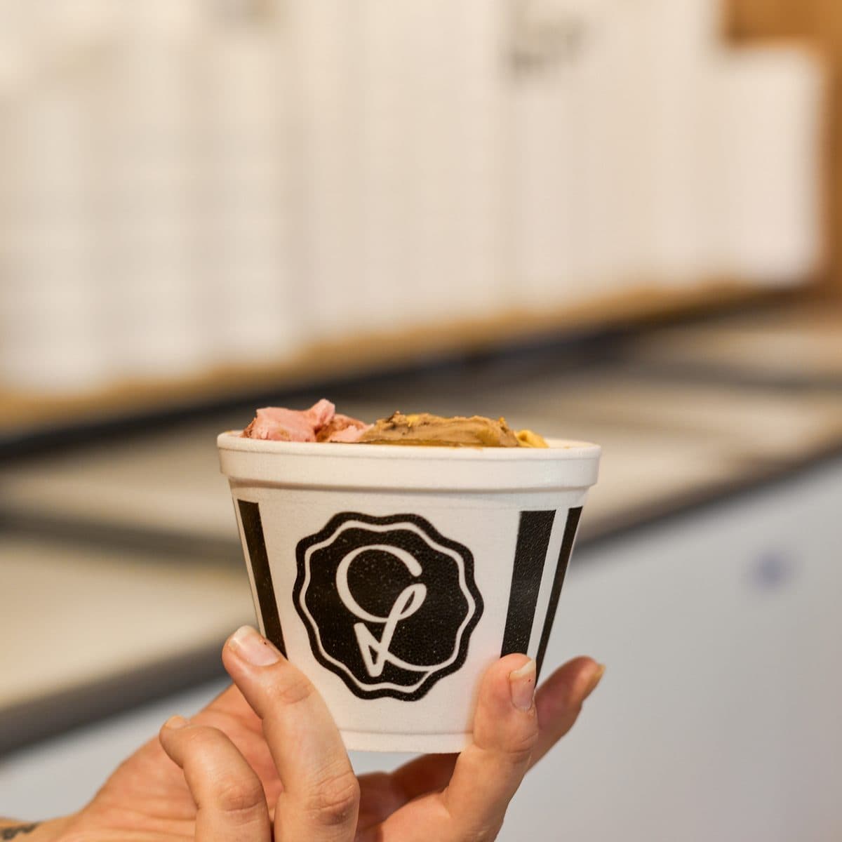 Una mano sosteniendo un potecito de helado con el sello de Libereco, con dos sabores adentro y las pilas de vasitos desenfocadas al fondo