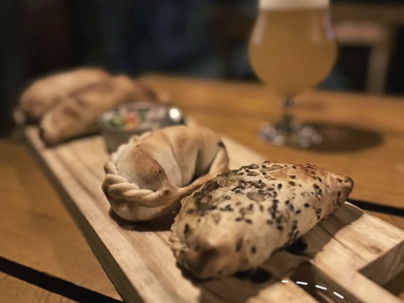 Dos empanadas sobre una tabla de madera, una con semillas de sésamo negro por encima, y una copa de cerveza detrás