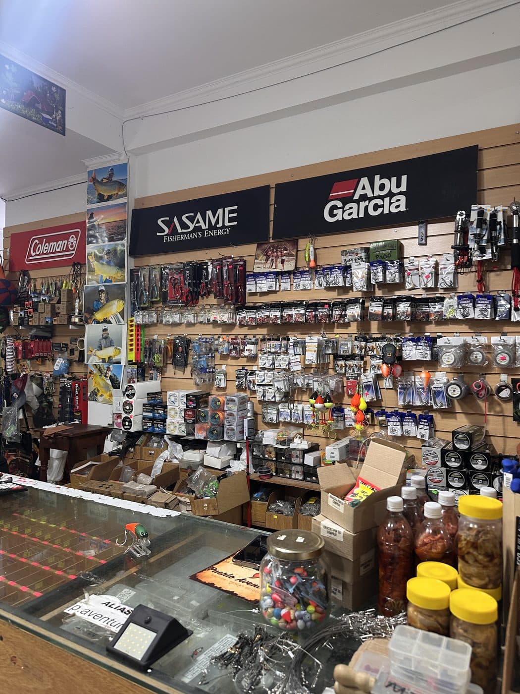 La pared de aparejos: anzuelos, plomadas y señuelos bajo los carteles de Abu Garcia y Sasame, con los frascos de carnada sobre el mostrador
